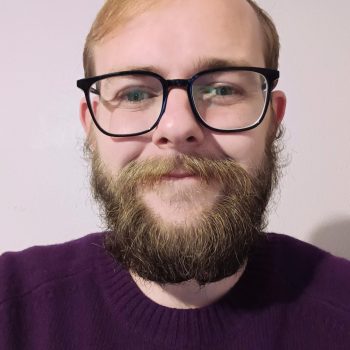 the photo shows our staff member Alex smiling straight at the camera. He is wearing glasses and has short brown hair and a big beard