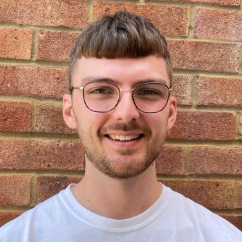 the photo shows our staff member Joe standing outside in front of a brick wall and smiling straight at the camera, he has dark eyes, glasses and short brown hair with a slight beard