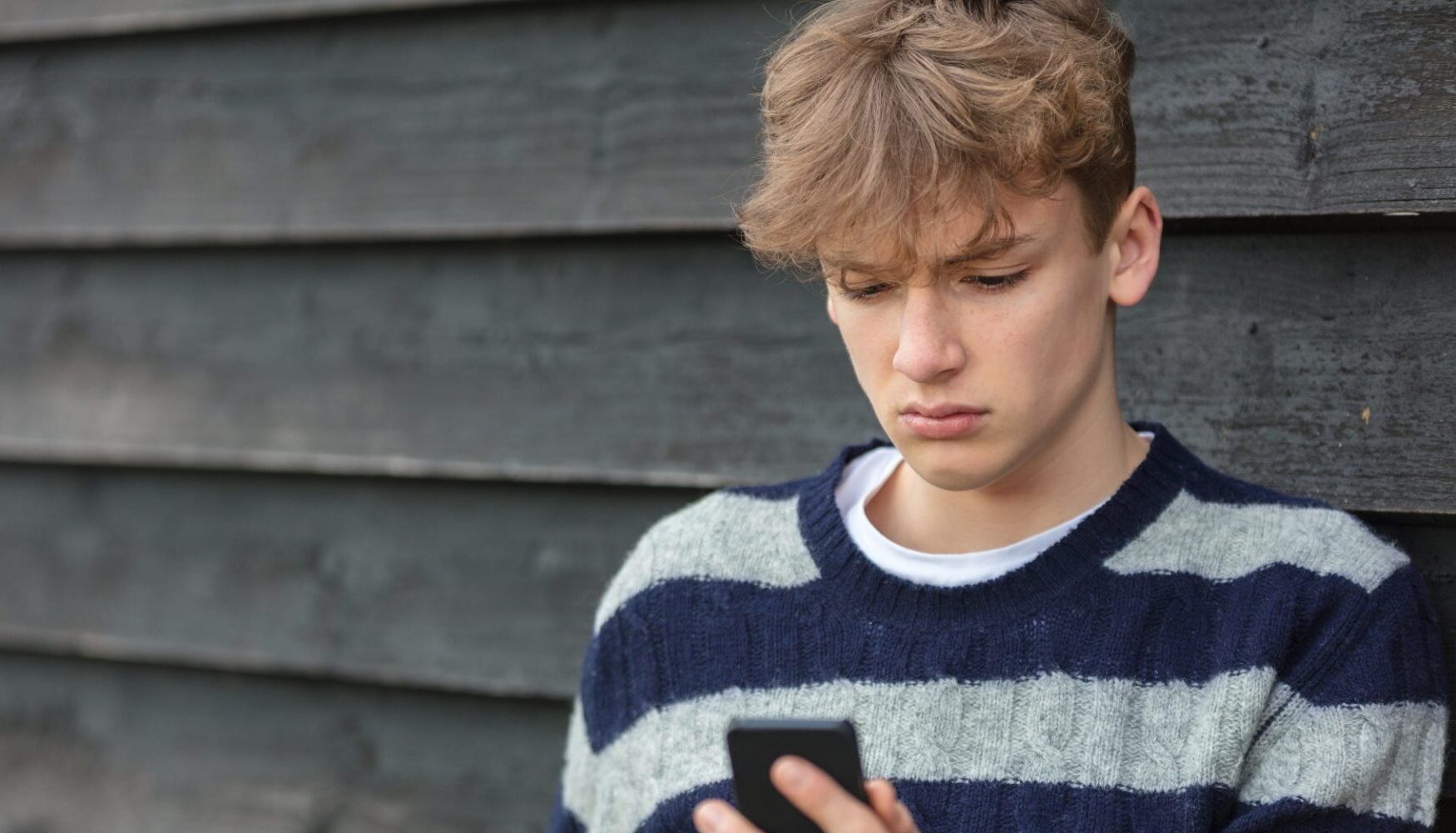 a teenage boy with blond hair is looking straight at the mobile phone in his hand with a confused expression