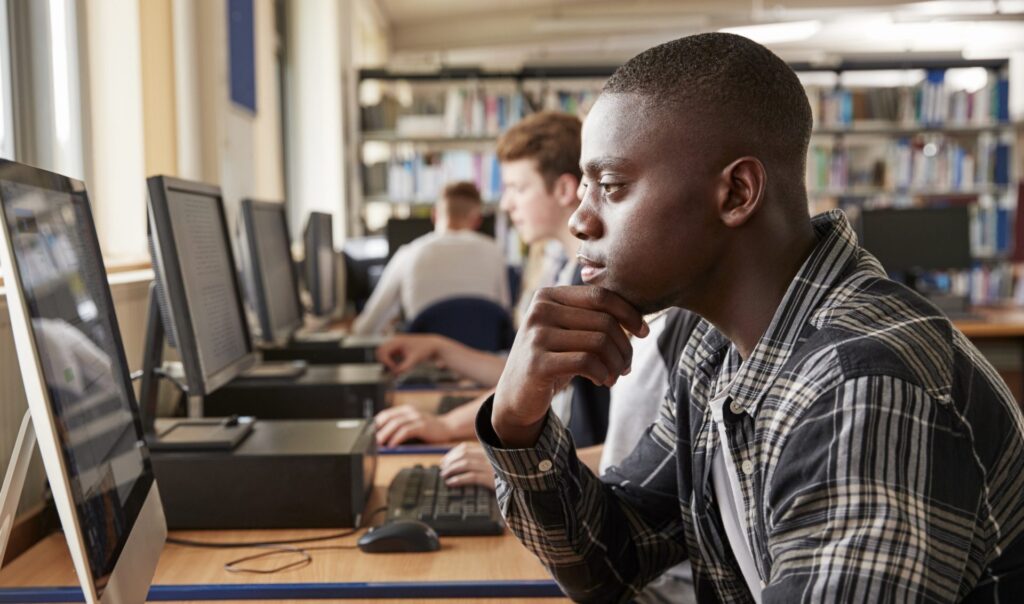 a teenage boy is sitting at a desk and looking at a computer monitor with a neutral expression