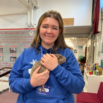 the photo shows our staff member Talitha standing inside and holding a guinea pig while smiling at the camera, she has dark eyes and straight brown shoulder length hair