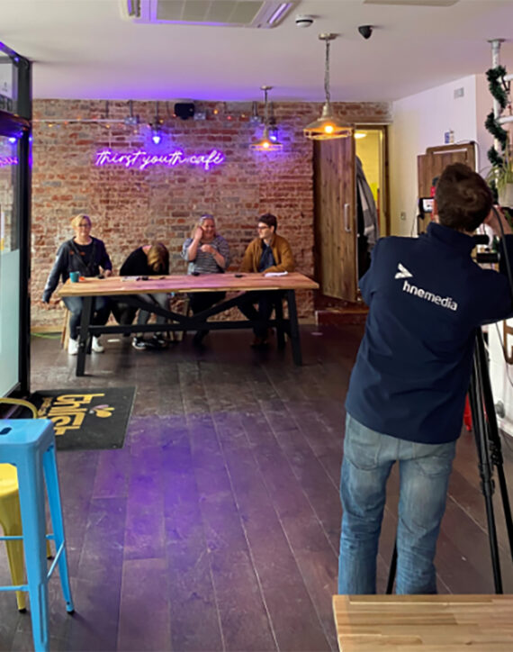 the image shows four people sitting down inside a cafe, with a camera man pointing a camera at them which is on a tripod