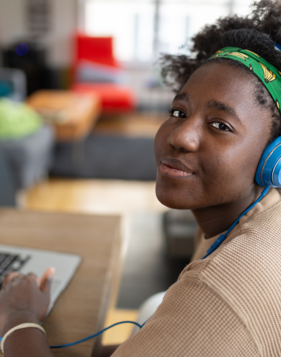 the photo shows a teenage girl wearing blue headphones and smiling straight at the camera, with one hand on her laptop