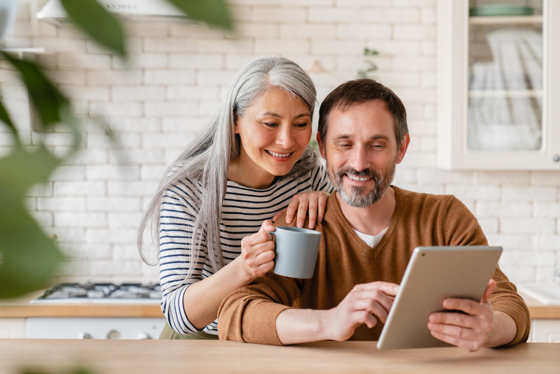 an older couple are indoors in a kitchen and are looking at a tablet computer together and smiling