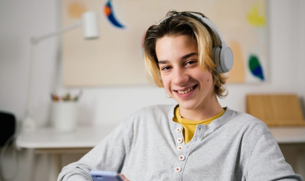 a teenage boy is wearing a grey pair of headphones and is sitting indoors holding his phone and smiling straight at the camera