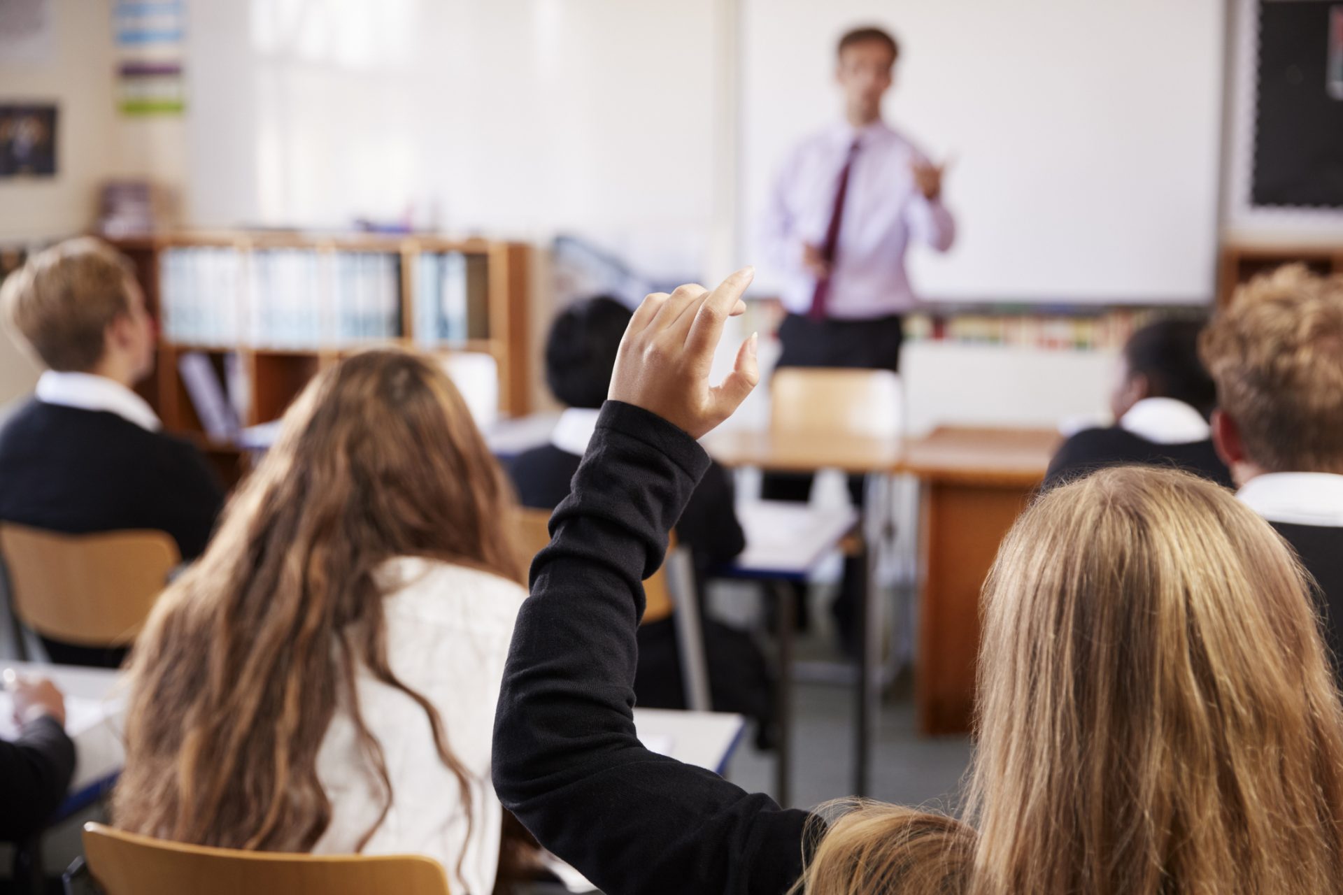 the photo shows a classroom is students from behind, with one girl raising her hand to the teacher at the front