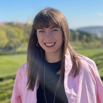 the photo shows our staff member Ilaria standing outside and smiling straight at the camera, she has dark eyes and long straight brown hair