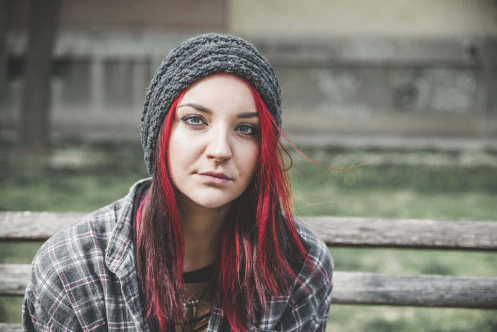 the photo shows a teenage girl with green eyes and long red hair sitting on an outside bench and looking straight at the camera with a neutral expression