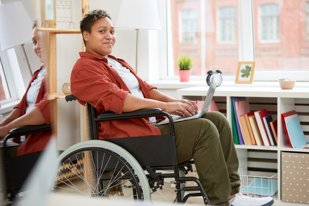 the photo shows a woman with short curly hair sitting in a wheelchair and smiling straight at the camera while holding her laptop computer