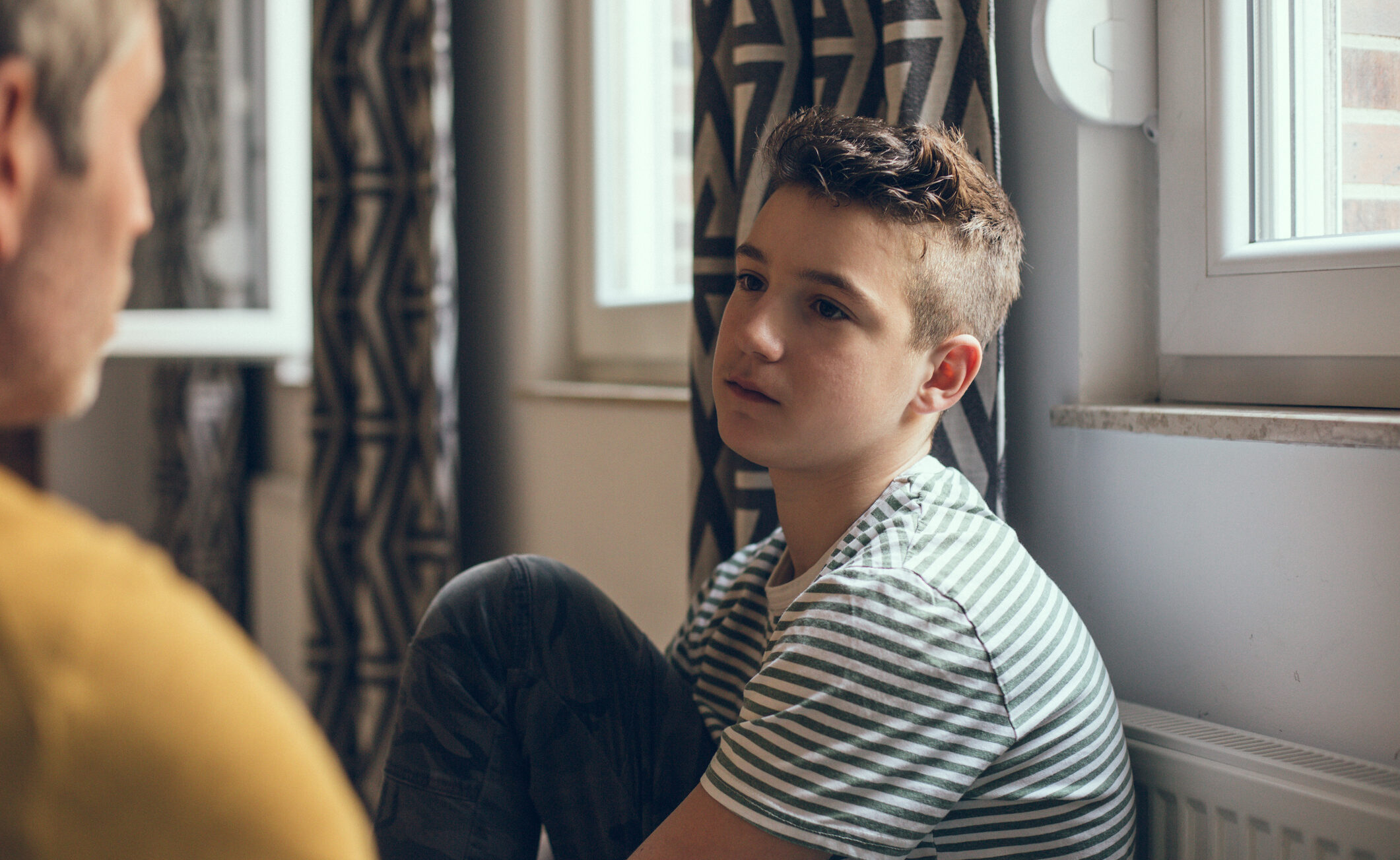 a teenage boy is sitting in a room against the wall and is looking at an adult with a serious expression