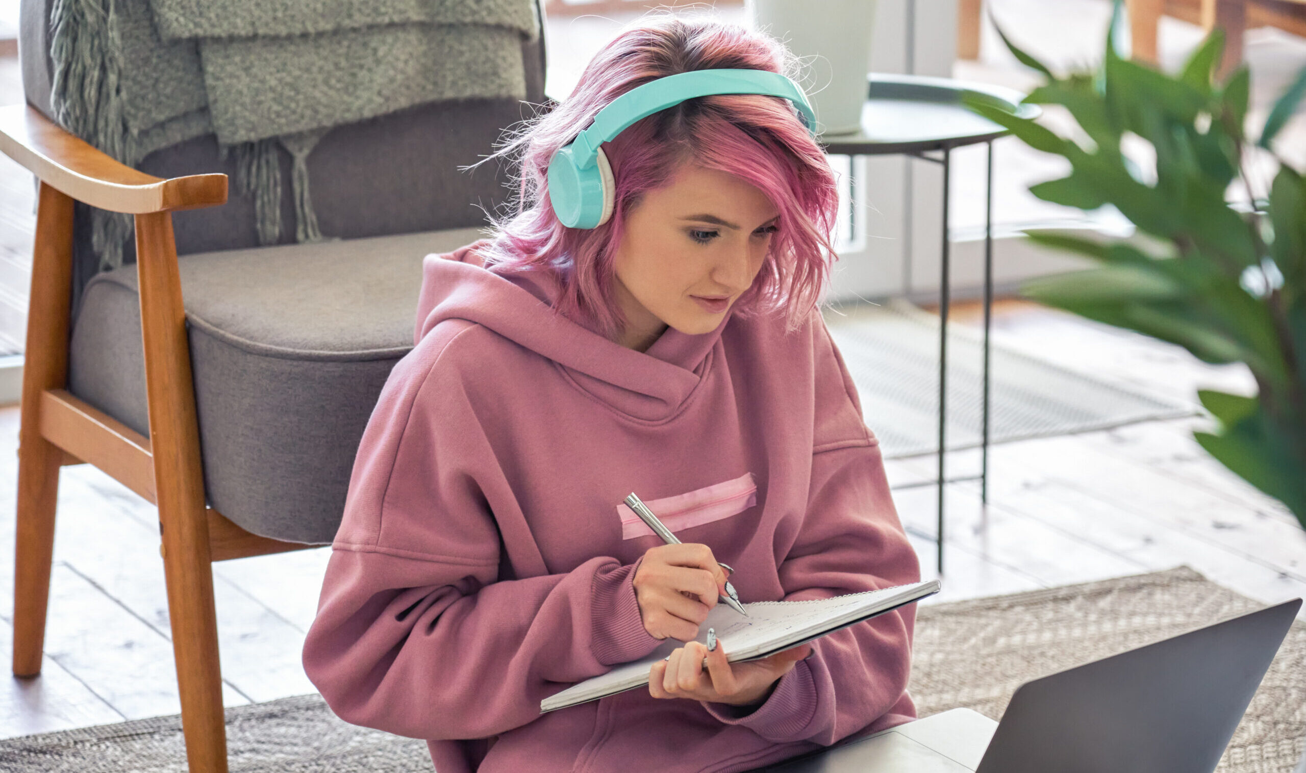 the photo shows a teenage girl with pink hair wearing blue headphones and a pink hoodie, she is holding a pen and textbook while looking at a laptop