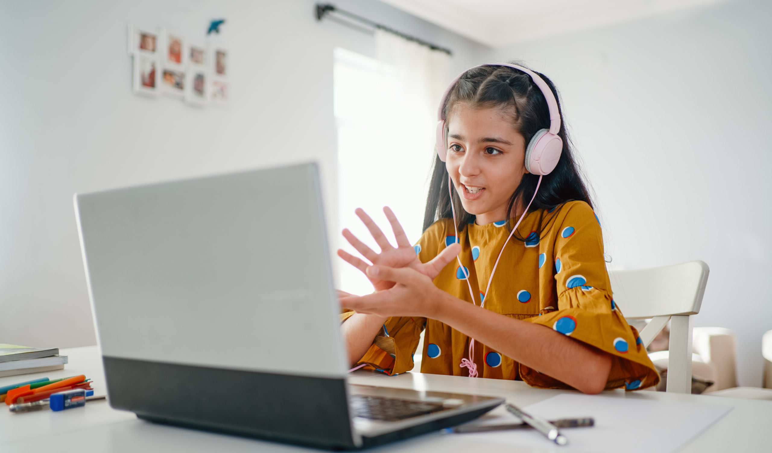 the photo shows a teenage girl wearing pink headphones and talking at her laptop screen