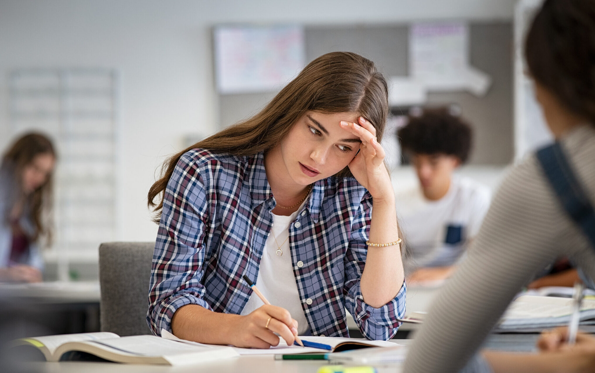 a teenage girl with long dark hair and dark eyes is sitting at a desk holding a pencil and looking downwards with a worried expression