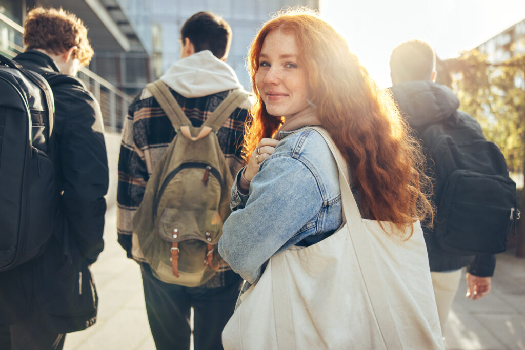 the photo shows the back of three teenage boys walking away, and one teenage girl turning around and smiling straight at the camera, she has long ginger hair and is holding a white tote bag