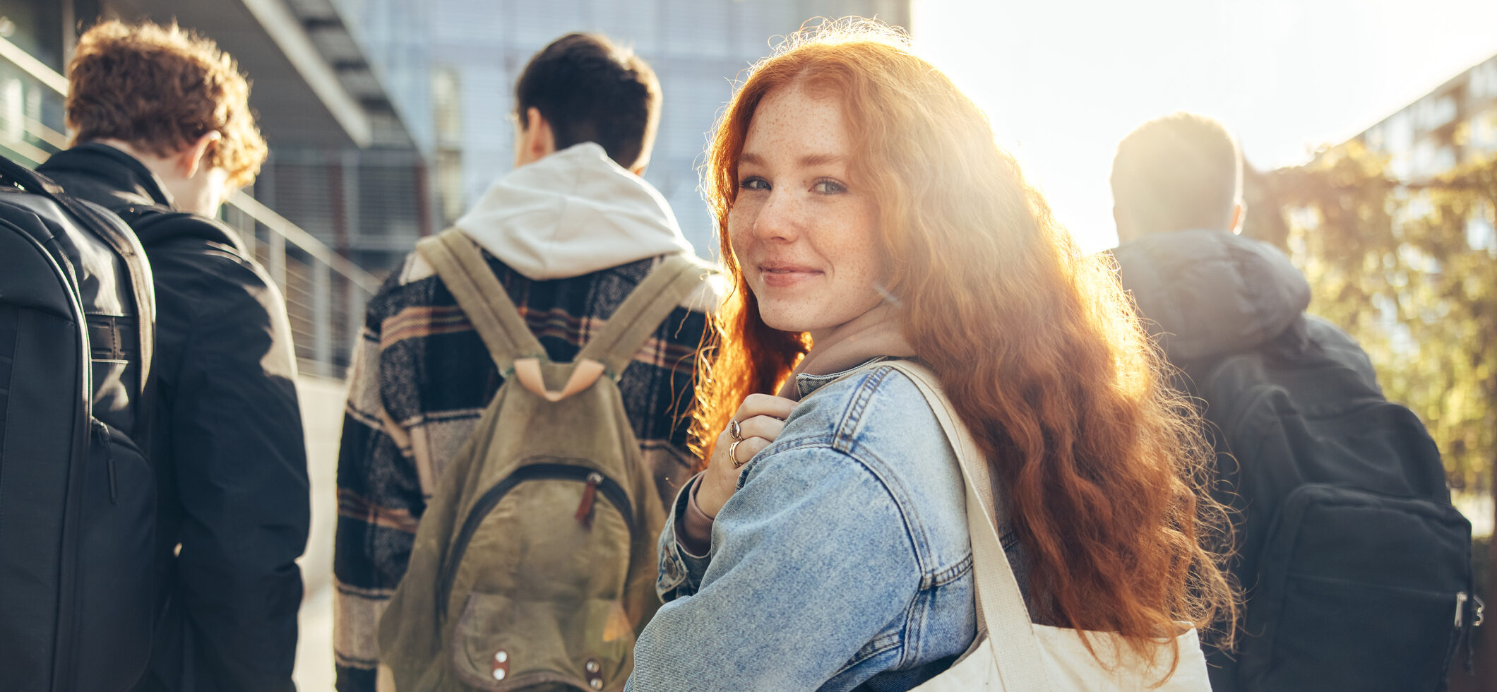 the photo shows the back of three teenage boys walking away, and one teenage girl turning around and smiling straight at the camera, she has long ginger hair and is holding a white tote bag