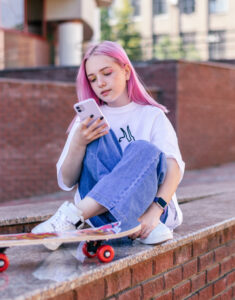 a young girl with long pink hair is sitting on a wall and looking down at her mobile phone with a skateboard next to her