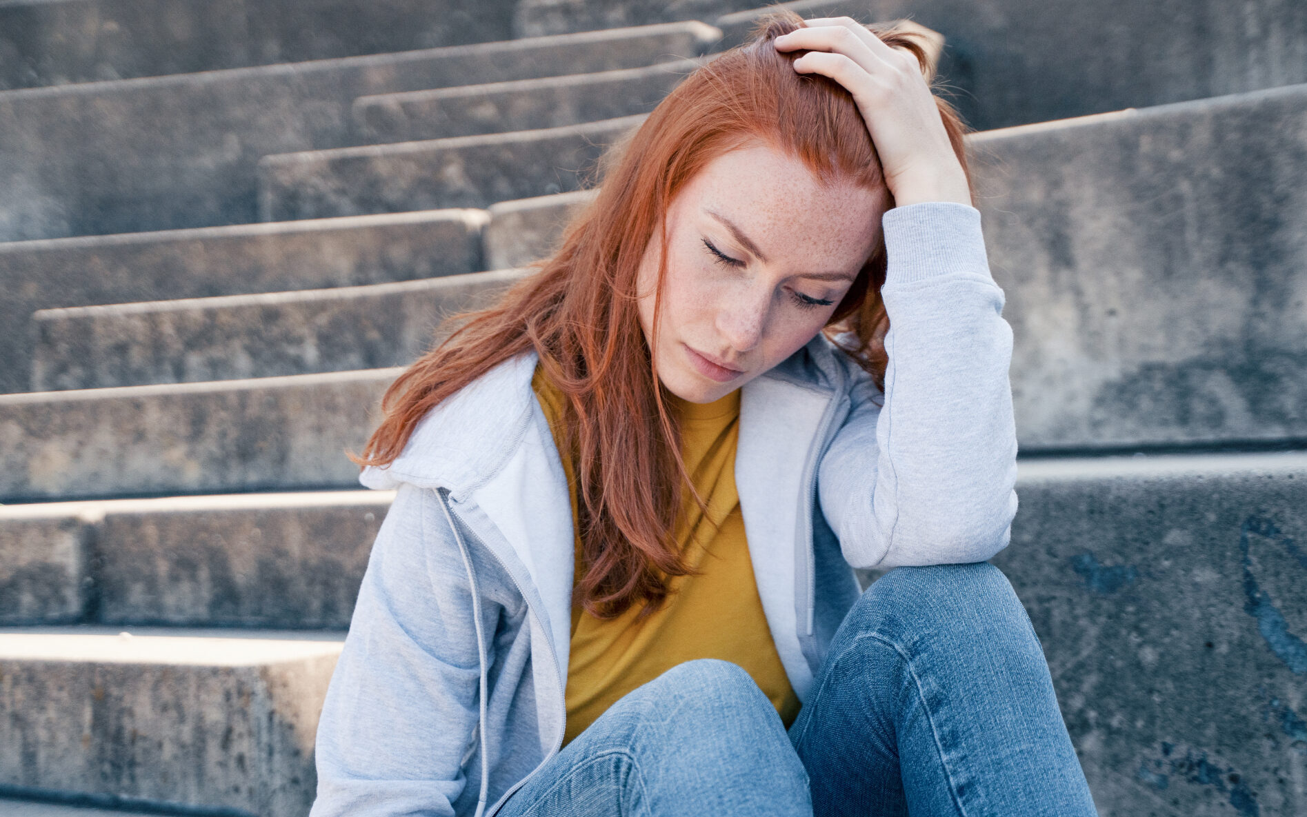 a teenage girl with long ginger hair is sitting down against some outside steps and looking downward toward the ground with a sad expression