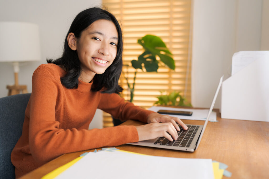 the photo shows a young girl with dark eyes and long black hair smiling straight at the camera with her hands on her laptop keyboard