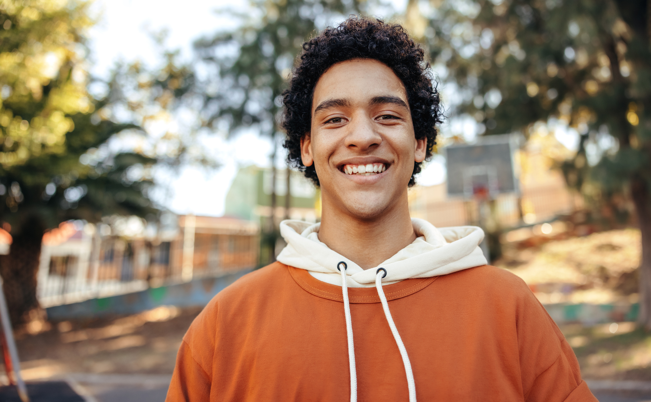a teenage male with black curly hair and dark eyes is standing outside and smiling straight at the camera
