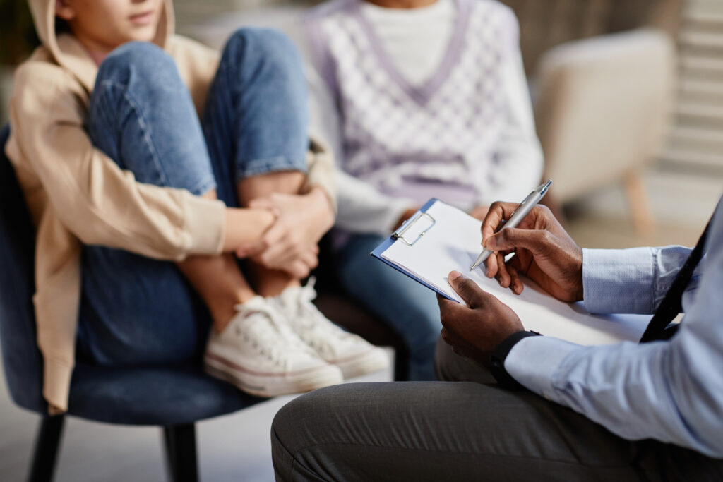 the photo shows an adult partially off screen holding a pen and clipboard, next to a young person sitting on a chair holding their legs against their body