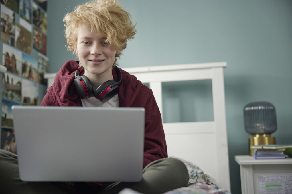 a teenage boy is wearing black headphones and is smiling straight at his laptop screen