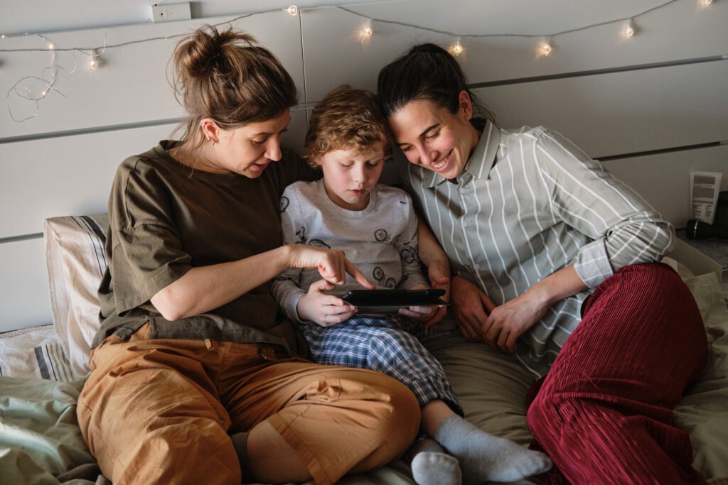 the photo shows two female adults sitting either side of a young boy in his pyjamas using a tablet in a large bed