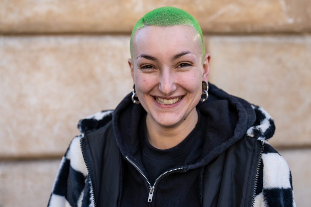 the photo shows a woman with short green hair and dark eyes who is outside and smiling straight at the camera