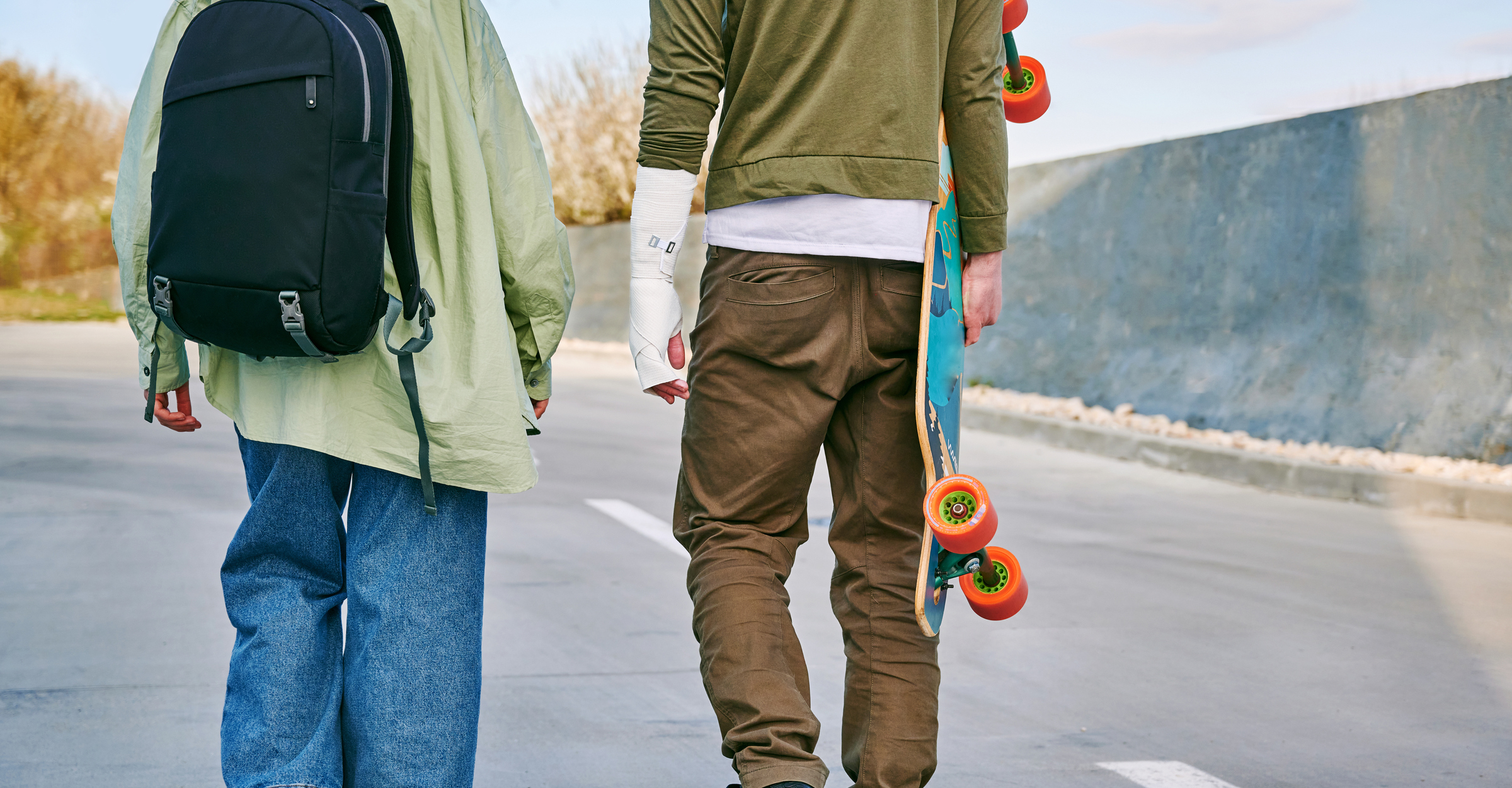 the backs of two teens are visible but not their heads, they are walking in a road and one of them is carrying a skateboard
