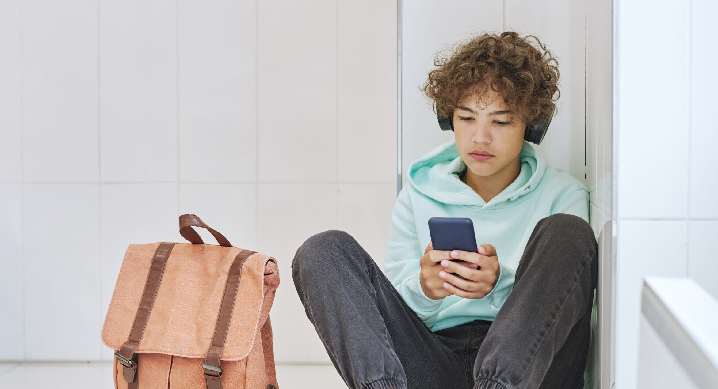 a teenage boy with brown curly hair is sitting down against a wall holding his phone is both hands and wearing headphones