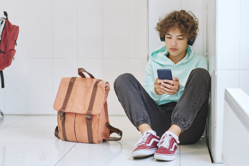 a teenage boy with brown curly hair is sitting down against a wall holding his phone is both hands and wearing headphones
