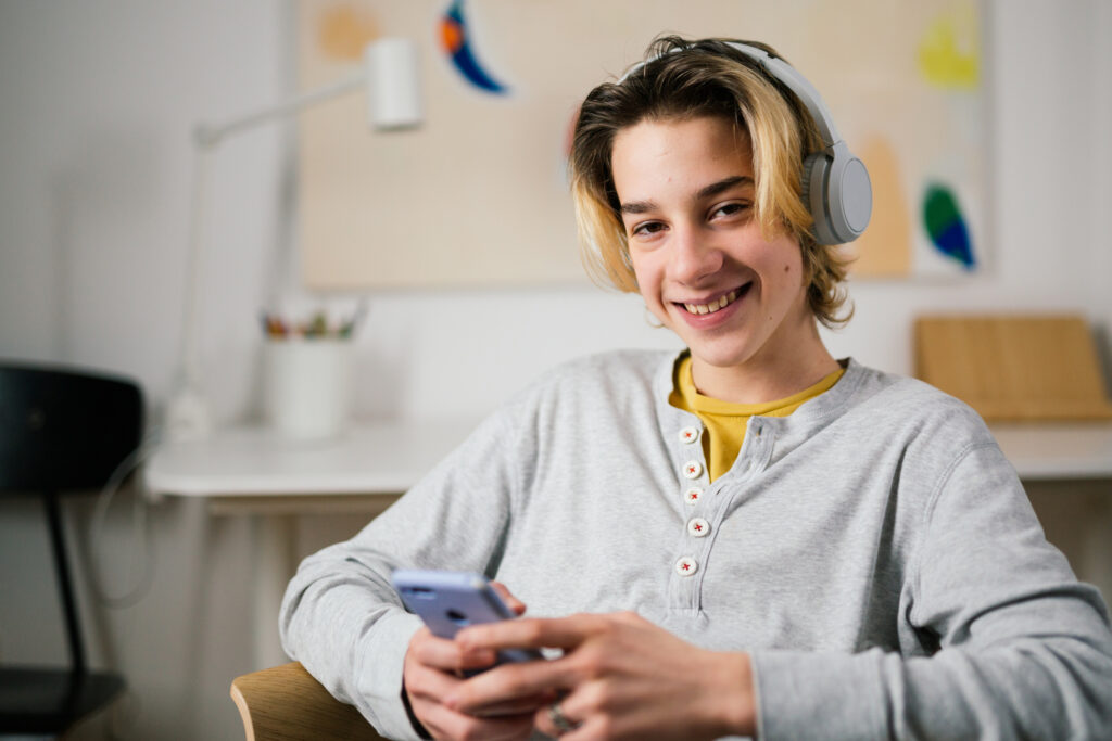 a teenage boy is wearing a grey pair of headphones and is sitting indoors holding his phone and smiling straight at the camera