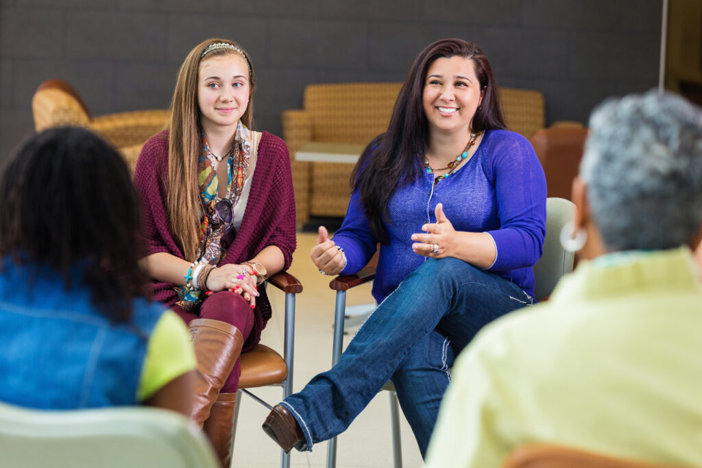 the photo shows a young girl sitting next to a female adult who is smiling at looking at someone opposite her in the group circle meeting