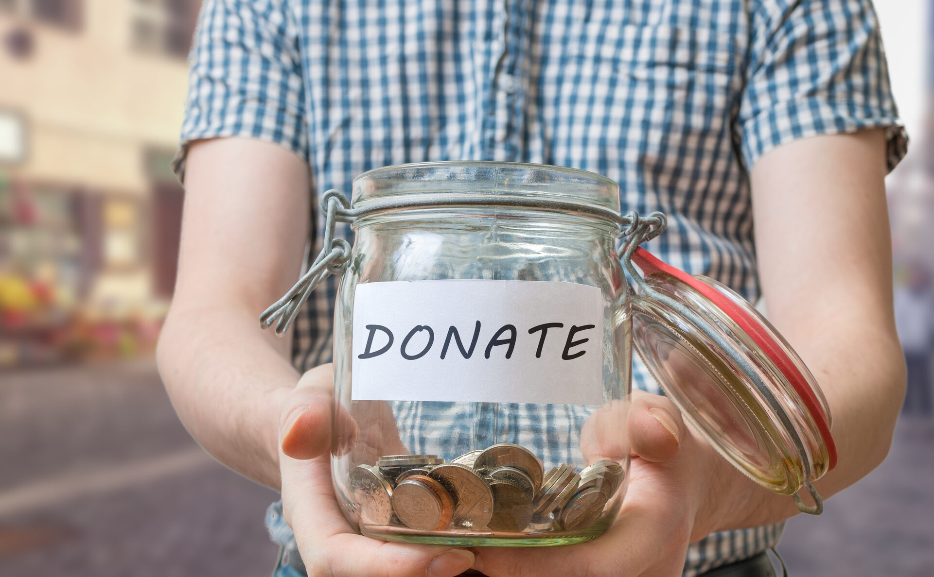 the photo shows the torso of a person who is holding a glass jar containing coins and has the word DONATE displayed on a white sticker