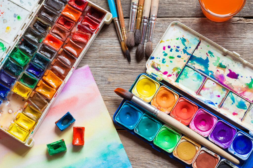 the photo shows a selection of watercolour paint and paint brushes on an old wooden background