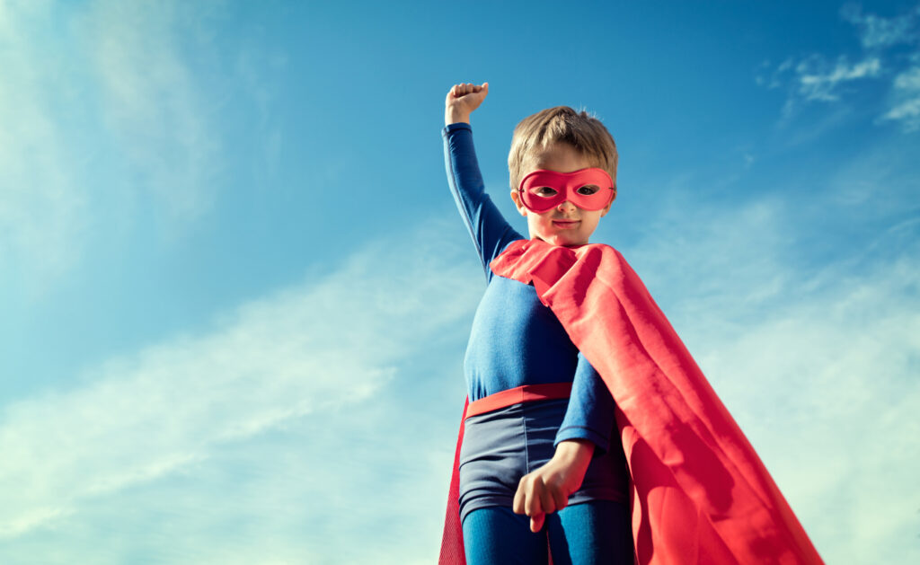 the photo shows a young boy who is dressed in a blue and red superhero costume with a blue sky background, he has one fist in the air and is smiling straight at the camera