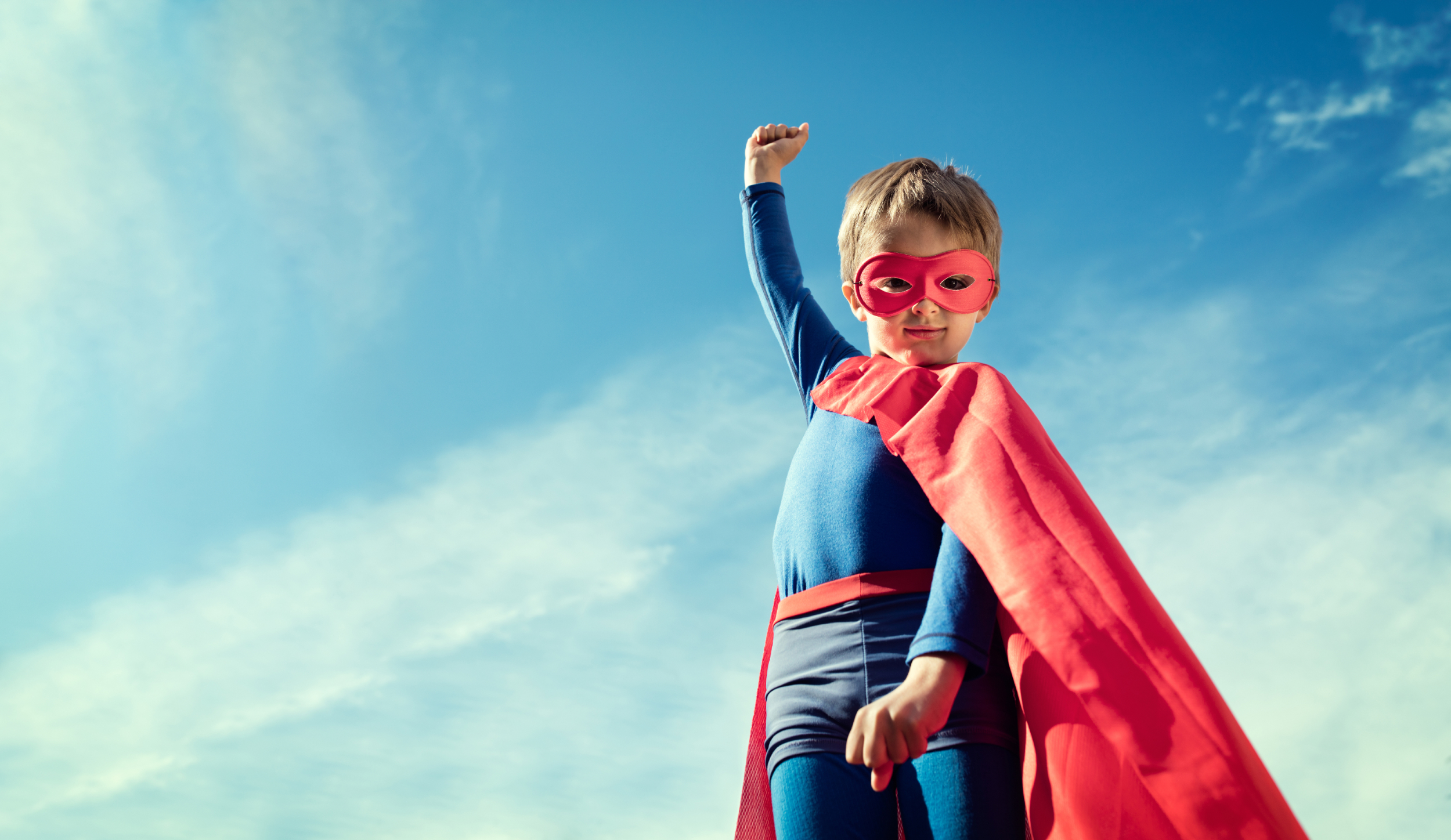 the photo shows a young boy who is dressed in a blue and red superhero costume with a blue sky background, he has one fist in the air and is smiling straight at the camera