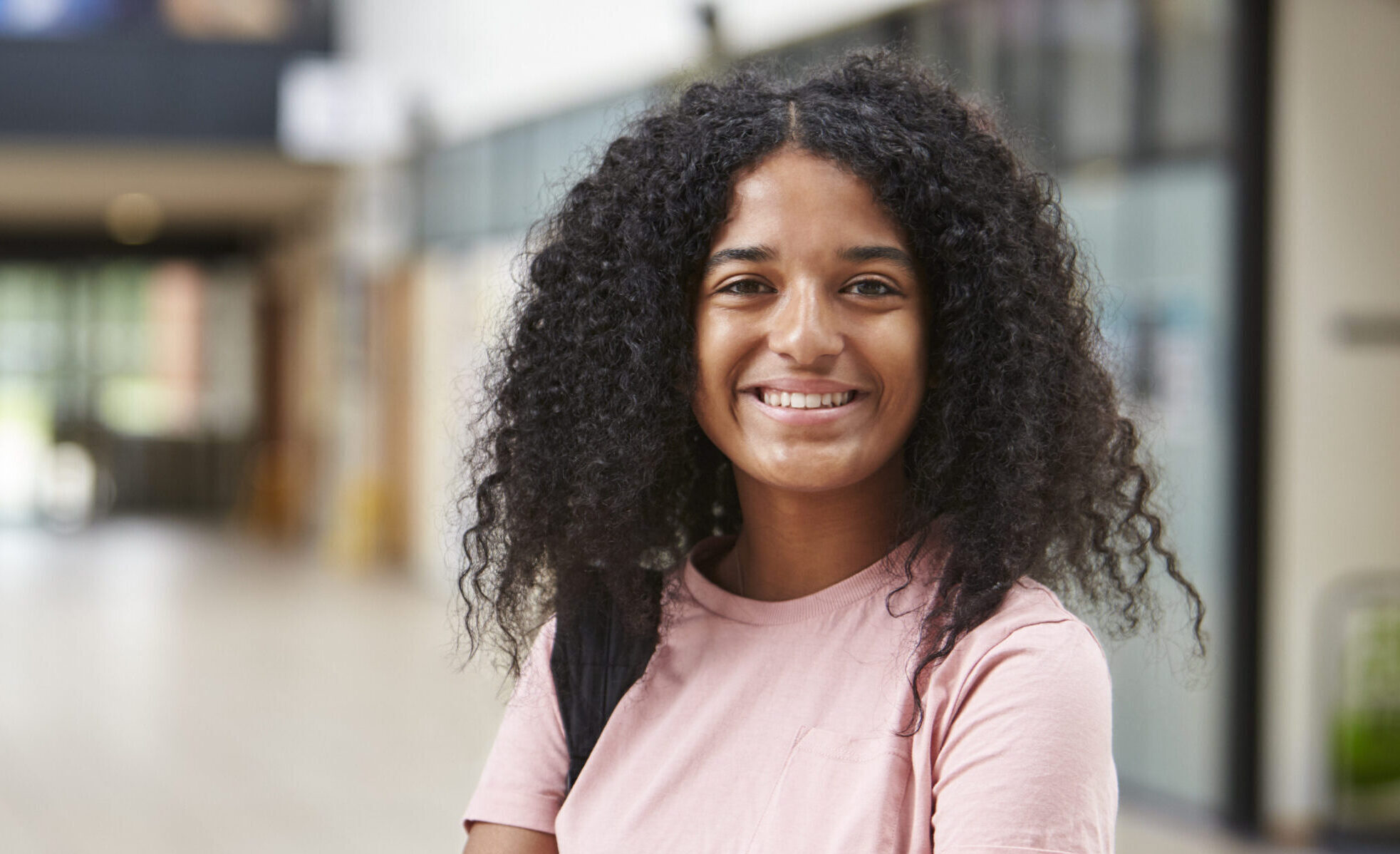 the photo shows a young female with long curly black hair and dark eyes, she is standing in a corridor and is smiling straight at the camera