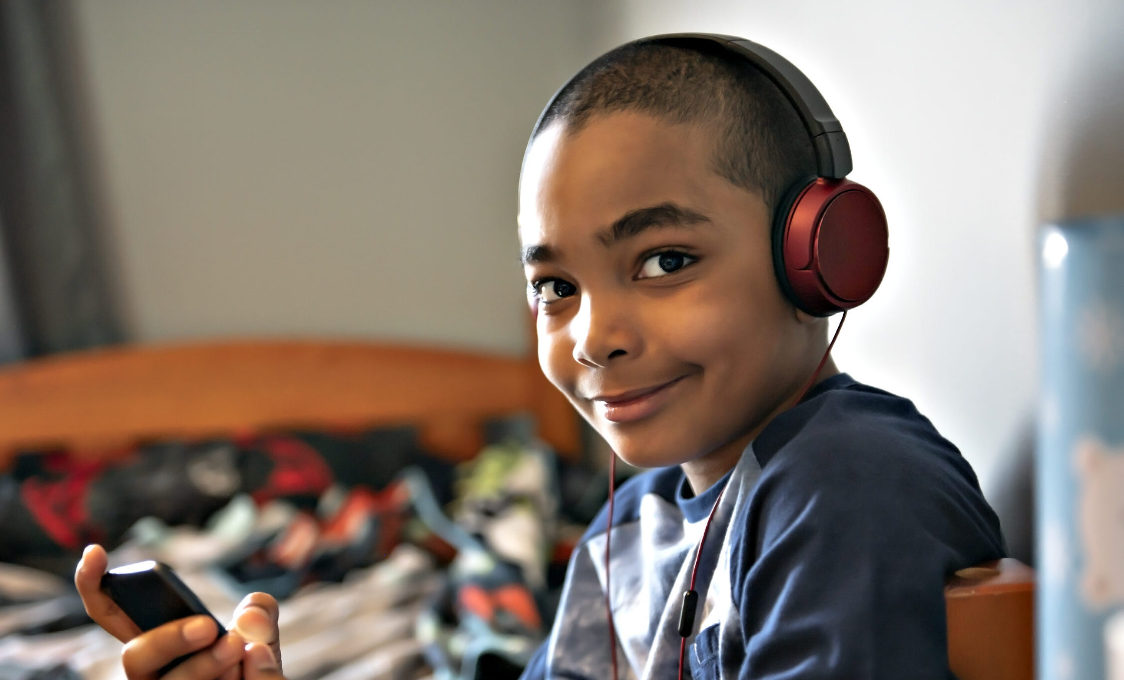 the photo shows a boy with short dark hair and eyes, who is sitting at the end of his bed wearing red headphones and smiling straight at the camera
