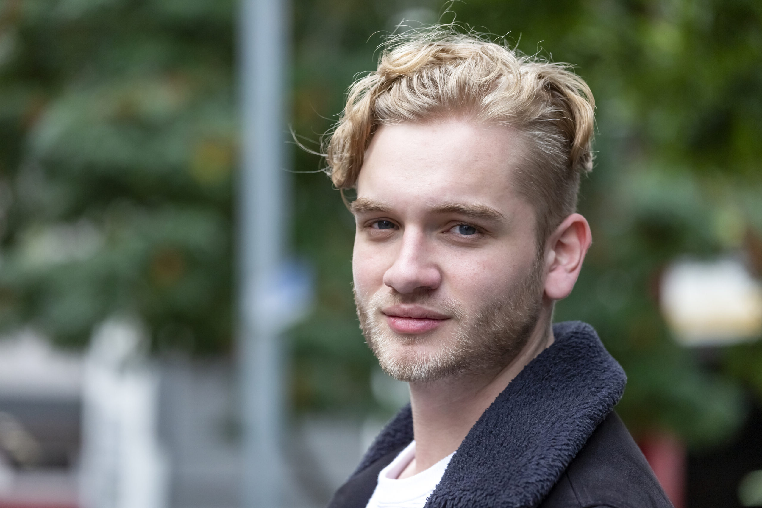 the photo shows a young man outdoors, who has blond hair and a trimmed beard, he is looking straight at the camera with a slight smile