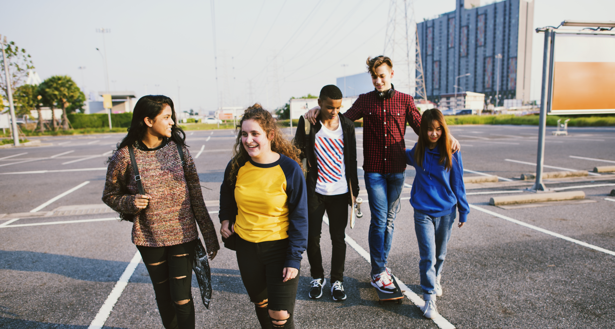 a group of mixed gender teenagers are walking across an outdoor car park and smiling at each other