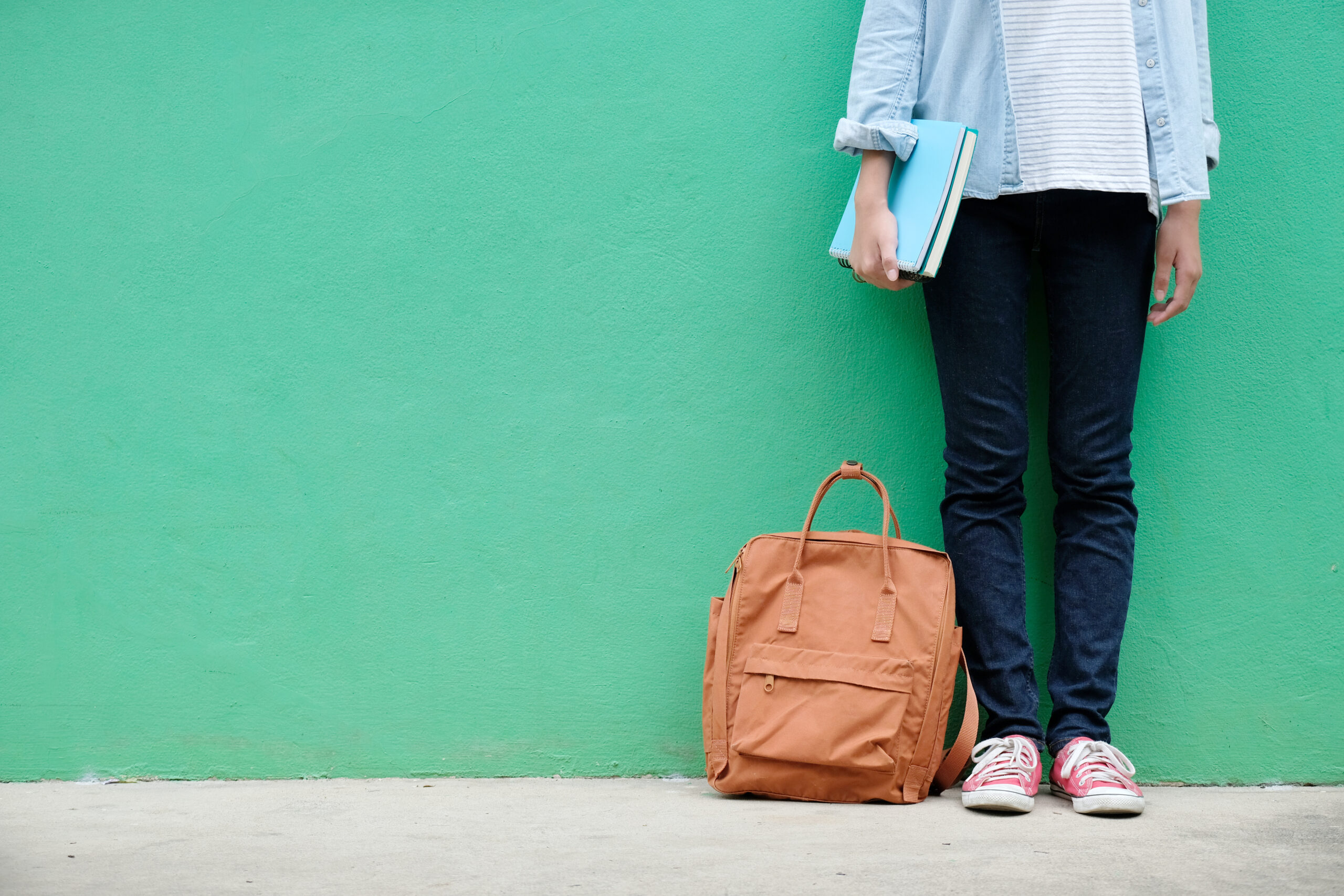 the image shows the bottom half of a young person standing and holding their books, with their bag by their feet