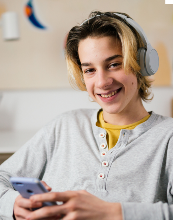 a teenage boy is wearing a grey pair of headphones and is sitting indoors holding his phone and smiling straight at the camera