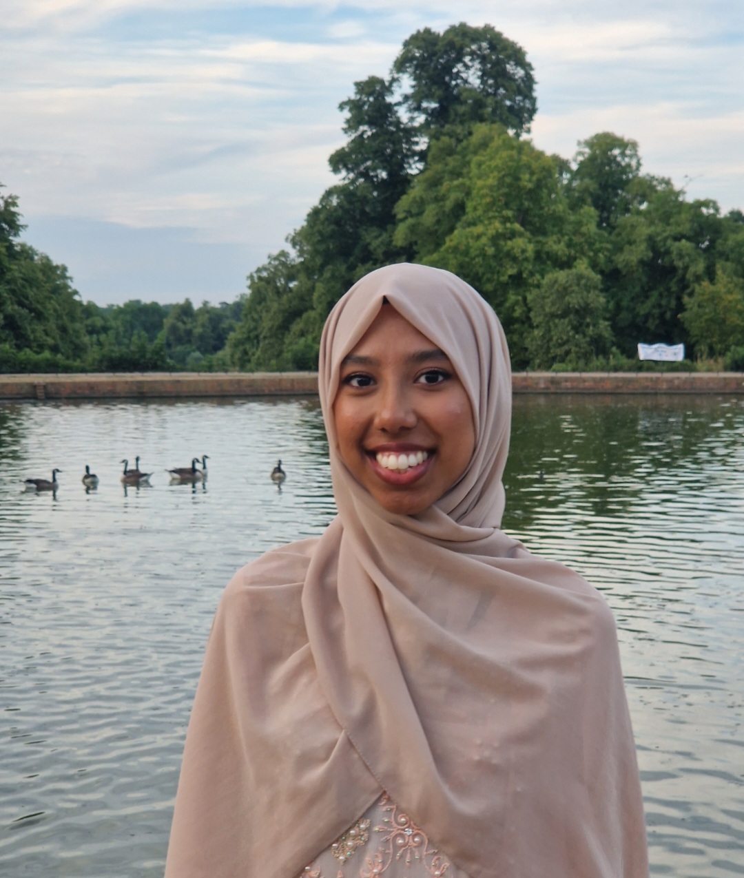 the photo shows our staff member Anjuma standing in front of an outdoor lake with ducks, she is smiling straight at the camera