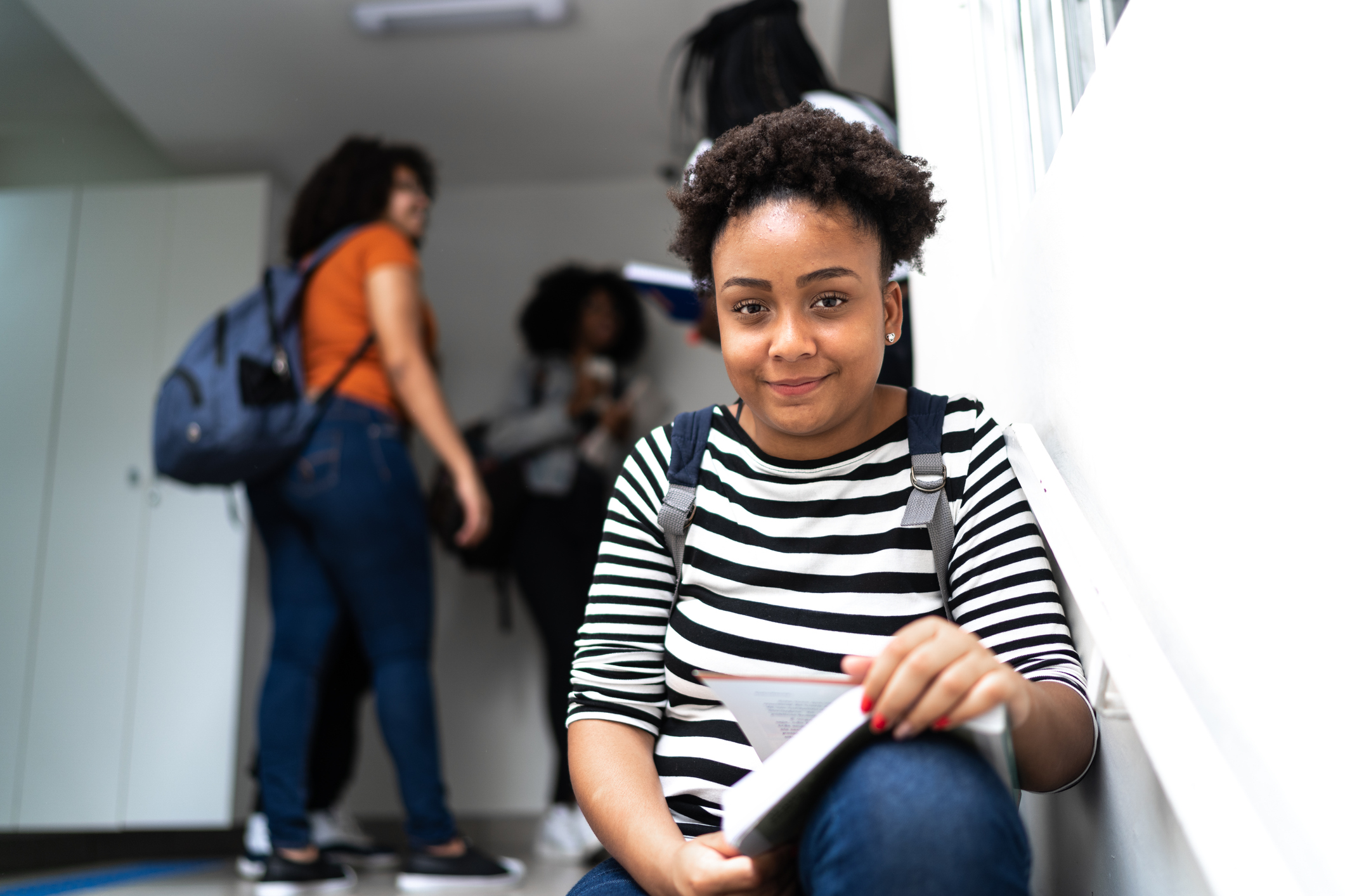 the photo shows a young girl with short afro hair, who is wearing a rucksack and sitting at the top of the school stairs holding a book and smiling straight at the camera