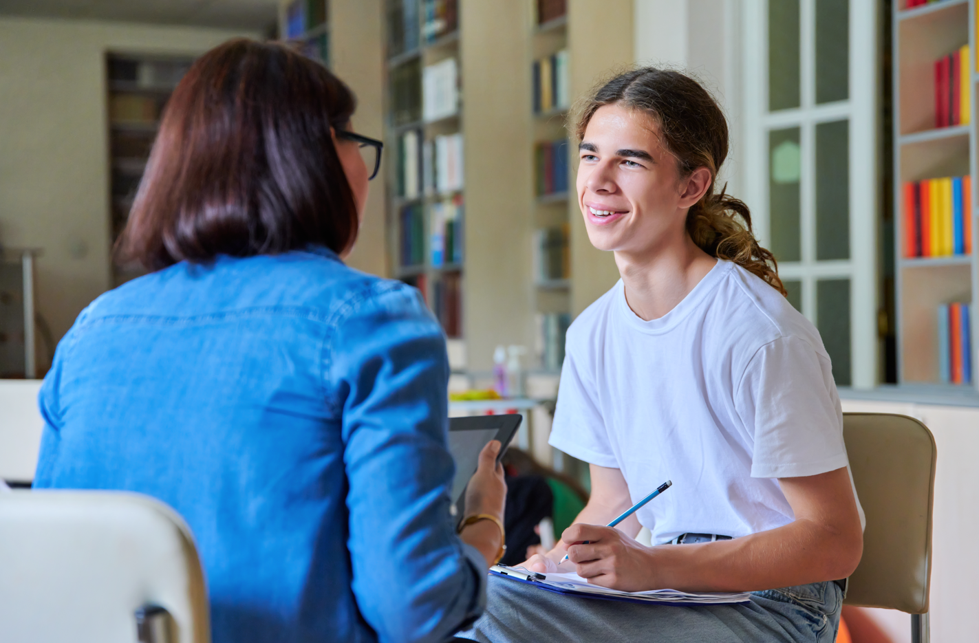 the photo shows a meeting between a female adult and a male young person, who is smiling at the adult and holding a pencil and clipboard