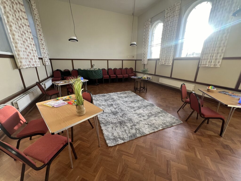 the photo shows a big empty room with tables and chairs spread out around a rug on the wooden floor, light is coming through the open curtains